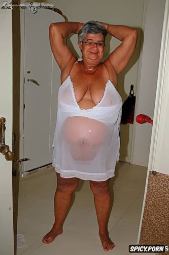 wearing a sleeveless white sheer night gown, a photo of a short ssbbw hispanic pregnant granny standing up in the badroom